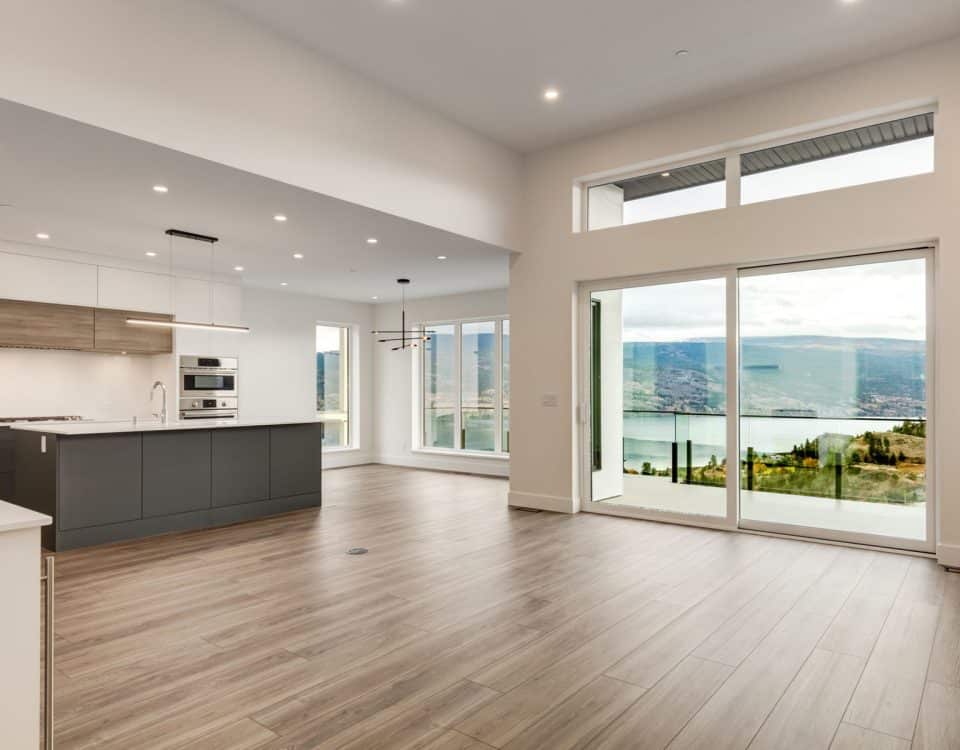 Spacious, modern kitchen and living area with floor-to-ceiling windows and glass doors, offering a panoramic view of a serene lake and mountains.
