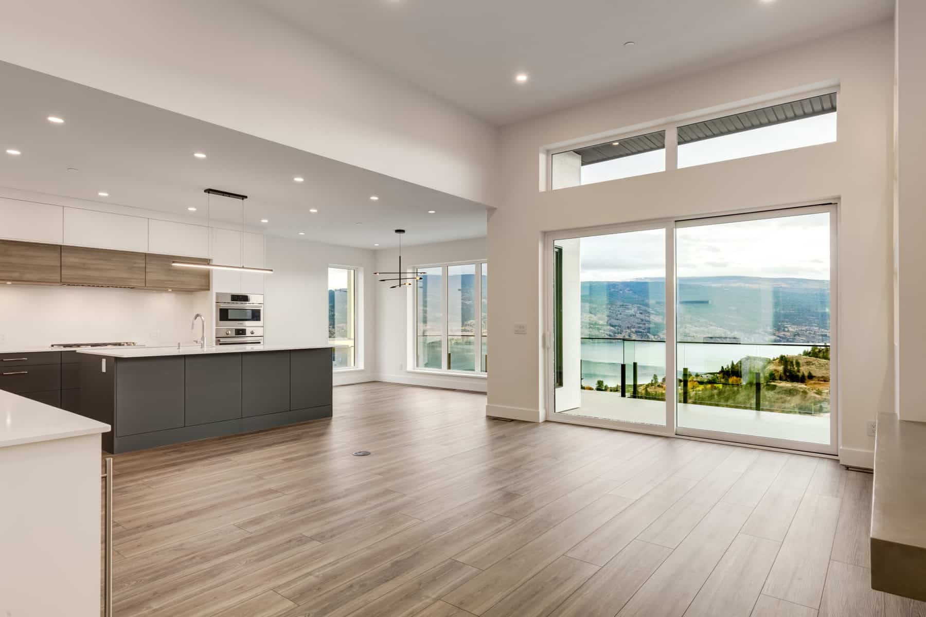 Spacious, modern kitchen and living area with floor-to-ceiling windows and glass doors, offering a panoramic view of a serene lake and mountains.