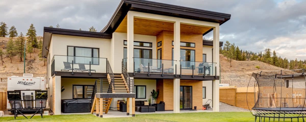 A modern home featuring a spacious yard and a large deck, surrounded by greenery and a clear blue sky.