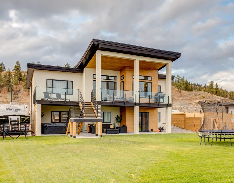 A modern home featuring a spacious yard and a large deck, surrounded by greenery and a clear blue sky.