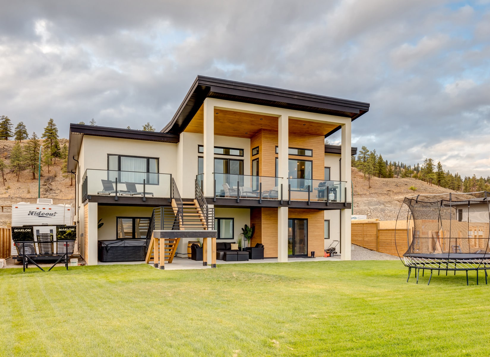 A modern home featuring a spacious yard and a large deck, surrounded by greenery and a clear blue sky.